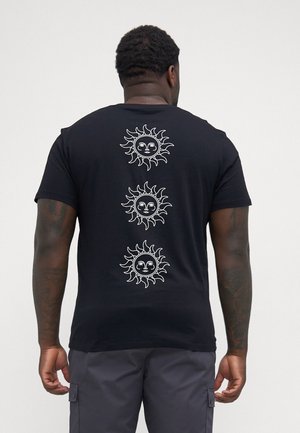 Mand iført sort T-shirt med tre hvide solansigter, der er lodret justeret på ryggen, kombineret med mørke cargobukser.