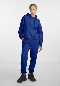 Modra kapuca s vrvico, v kombinaciji z usklajenimi trenirka hlačami. Obe sta narejeni iz mehke tkanine. Črne debele superge dopolnjujejo izgled.