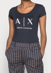 Camiseta de color azul marino de manga corta y escote redondeado, que presenta un logo blanco de "A|X Armani Exchange". Combinada con pantalones de tiro alto a rayas.