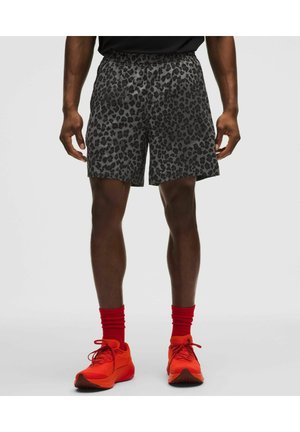 Mand iført grå og sort leopardmønstrede shorts, lyse røde sokker og matchende røde sportssko, stående på en ensfarvet lys baggrund.