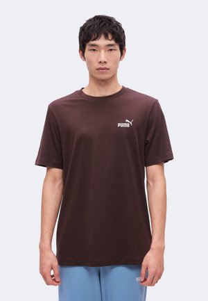 Ung mand iført en ensfarvet mørkebrun Puma t-shirt og lyseblå Puma træningsbukser, stående mod en ensfarvet lys baggrund.