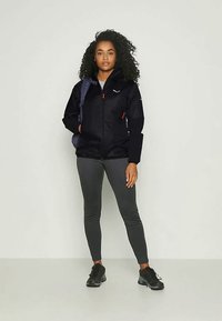 Giacca nera con zip e dettagli rossi, abbinata a leggings grigi e sneakers neri. Il modello porta anche uno zaino blu.
