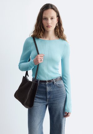 Jeune femme portant un pull côtelé bleu clair et un jean bleu, tenant un sac à épaule noir sur fond blanc uni.