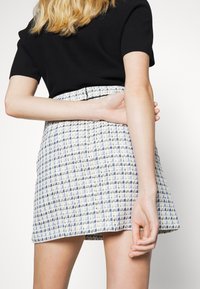 Mujer vistiendo una blusa negra de manga corta y una falda a cuadros azul y blanca, vista desde atrás con un brazo flexionado detrás de ella.