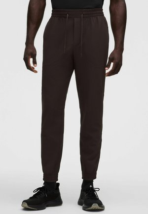 Man draagt zwarte joggerbroek met structuur, elastische boorden en aantrekkoord in de taille, gecombineerd met zwarte sportschoenen en sokken.