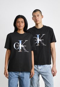 Crni pamučni majici s velikim grafičkim logom "CK" i "Calvin Klein Jeans" u višebojnom dizajnu, u kombinaciji s laganim trapericama.