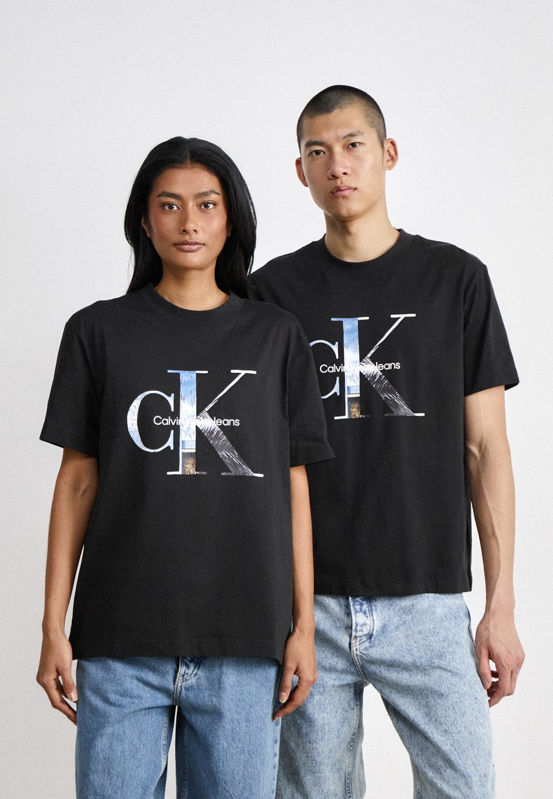 Crni pamučni majici s velikim grafičkim logom "CK" i "Calvin Klein Jeans" u višebojnom dizajnu, u kombinaciji s laganim trapericama.