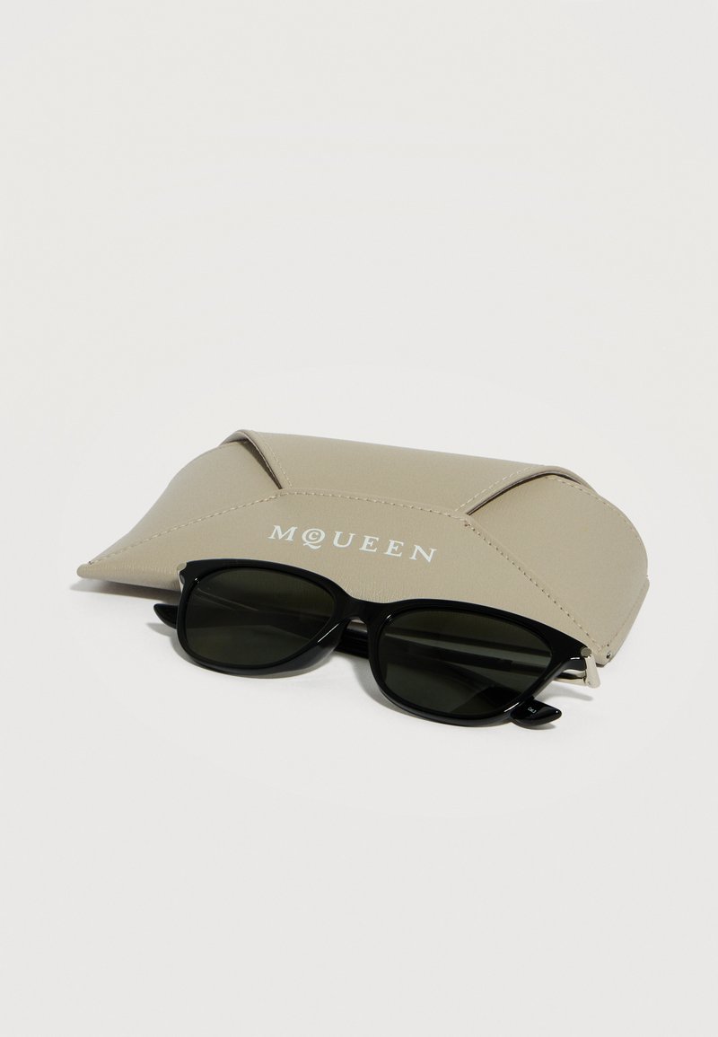 Black rectangular sunglasses resting in front of a beige McQueen branded leather case on a light background.