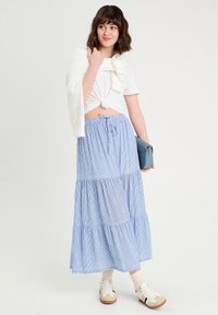 Jupe en tiered bleu et blanc à rayures, t-shirt blanc noué, pull blanc drapé sur les épaules, tenant un sac bleu clair, baskets décontractées.