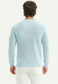 Pull en tricot bleu clair à manches longues, avec des poignets et un ourlet côtelés, présentant un motif texturé. Porté avec un pantalon blanc.