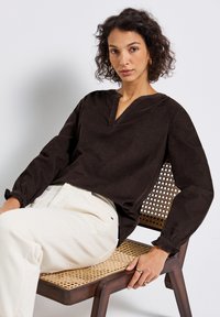 Vrouw met krullend haar, gekleed in een zwarte blouse met lange mouwen en witte broeken, zittend op een houten stoel met een geweven zitting, kijkend naar de camera.