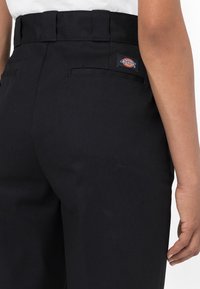 Svarta byxor med hög midja, med en bakficka, slät textur och en liten färgad logotyp på höger sida.