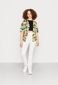 Camisa preta de mangas curtas com padrões verdes, laranjas e bege sobre um top cropped preto, combinada com calças flare brancas texturizadas e sapatos brancos.