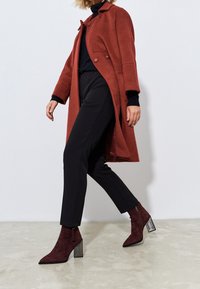 Mujer caminando vestida con un abrigo color óxido, pantalones negros, blusa negra y botas de ante burdeos con tacones de bloque plateados.