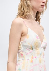 Νεαρή γυναίκα με κυματιστά ξανθά μαλλιά που φοράει λευκό φόρεμα με λεπτές τιράντες και παστέλ λουλουδάτα και γεωμετρικά σχέδια.