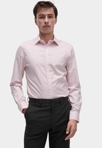 Camicia button-up rosa chiaro con colletto classico e maniche lunghe, abbinata a pantaloni grigi scuri, mostrata in vista frontale.