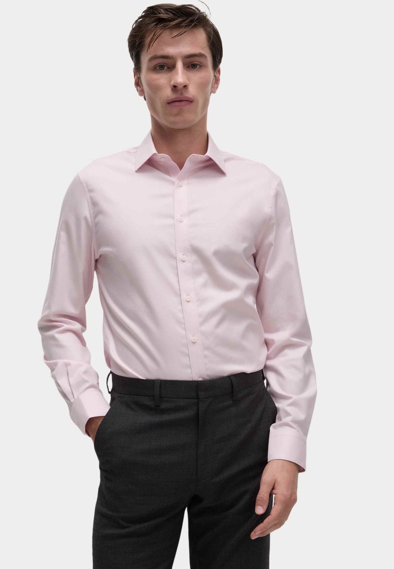 Camicia button-up rosa chiaro con colletto classico e maniche lunghe, abbinata a pantaloni grigi scuri, mostrata in vista frontale.