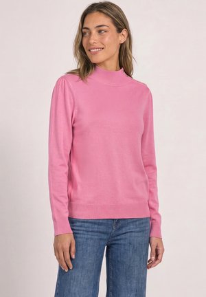 Femme aux cheveux mi-longs portant un pull rose à manches longues avec des boutons sur l'épaule et un jean bleu, souriante et regardant vers la gauche.