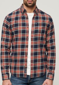Camisa de cuadros en azul marino con patrones rojos y blancos, de botón, en tejido de algodón, de manga larga, con un bolsillo en el pecho, superpuesta sobre una camiseta blanca.