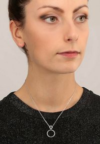 Sølv halskæde med en cirkulær pendants, der har en delikat kæde og en tekstureret sort sweater som baggrund.