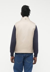 Bež prsluk s visokim ovratnikom, tamnoplavim dugim rukavima i glatkom teksturom. Nošen preko plavih traperica, s naglaskom na minimalistički dizajn.