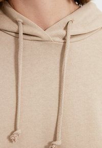 Sudadera beige con cuello cosido, cordones trenzados y material de algodón texturizado, destacando un diseño simple y limpio.