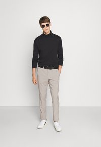 Jersey negro de cuello alto, pantalones beige claros y zapatillas blancas con detalles negros. Modelo con gafas de sol redondas oscuras. Fondo sencillo.