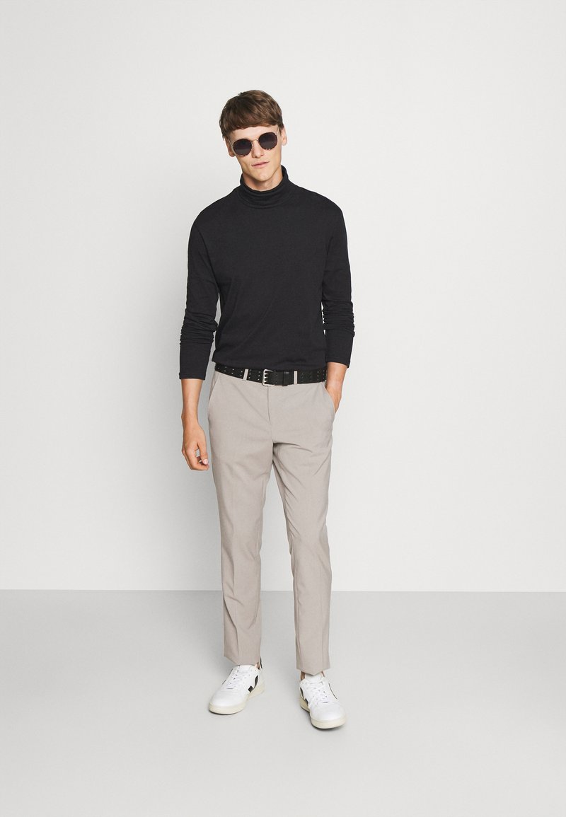Jersey negro de cuello alto, pantalones beige claros y zapatillas blancas con detalles negros. Modelo con gafas de sol redondas oscuras. Fondo sencillo.