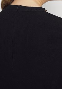 Haut en tissu côtelé noir avec un col montant et un petit zip à l'arrière. La texture est douce et ajustée.