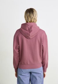 Burgunderfarget hettegenser med en cropped design, ribbestrikkede mansjetter og nederkant. Myk tekstur, vist på en person fra ryggen. Lys bakgrunn.
