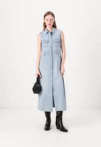 Lys blå denim kjole uten ermer med knappelukking, to brystlommer og en frontsplitt. Brukt med knelangne svarte støvler og en liten håndveske.