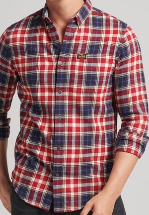 Mężczyzna w czerwono-niebiesko-białej kraciastej koszuli z podwiniętymi rękawami i małą kieszonką na piersi.