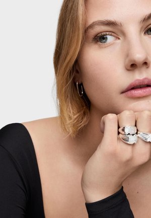 Drei silberne Ringe an einer Hand mit einem strukturierten Design und einem undurchsichtigen Stein in der Mitte. Das Modell hat schulterlanges blondes Haar vor hellem Hintergrund.