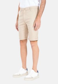 Pantaloni corti beige in cotone, con chiusura a bottone, orli arrotolati e tasche laterali, abbinati a sneakers bianche con accenti grigi.