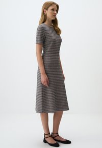 Robe mi-longue en tissu texturé à carreaux avec des manches courtes. Les couleurs comprennent le gris, le noir et des touches de rouge. Portée avec des ballerines noires.