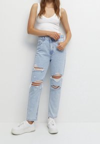 Jeans strappati blu chiaro con bordi sfrangiati e una vestibilità comoda. Abbinati a sneakers bianche. Presentano una vita alta e molte aperture danneggiate.