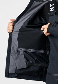 Personne tenant une veste noire ouverte montrant une grande poche intérieure en mesh avec le logo "Swedemount" sur la doublure.