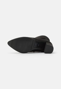 Bota de ante negra con una punta afilada y un tacón bajo y cuadrado. La suela es lisa y plana, con detalles texturizados sutiles.