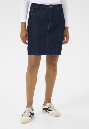 Personne portant une jupe en jean bleu foncé longueur genou, un haut blanc à manches longues et des baskets blanches avec des détails noirs, debout sur un fond blanc.