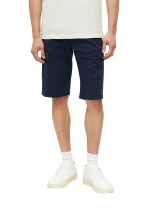 Homme portant un short cargo bleu marine, des chaussettes blanches, des baskets blanches et un t-shirt blanc, debout devant un fond blanc.