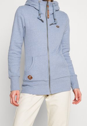 Sudadera con capucha azul claro con cremallera, capucha con cordón, bolsillos frontales y parches marrones con marca, combinada con pantalones blanco hueso.