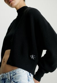 Černý pletený kardigan s texturovaným žebrováním, zapínáním na knoflíky a logem. Stylizovaný s přiléhavým černým topem a světlým denimem.