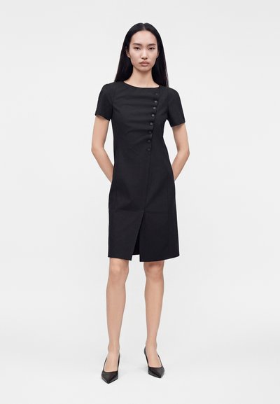 Femme portant une robe noire ajustée longueur genou avec manches courtes, détail des boutons sur le côté, fente à l’avant et escarpins noirs à bout pointu, debout, mains derrière.