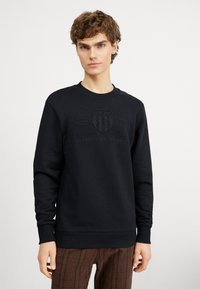 Fekete pulóver kerek nyakkal, kiemelkedő GANT logóval és "American Sportswear" felirattal. Simább, puha anyagból készült.