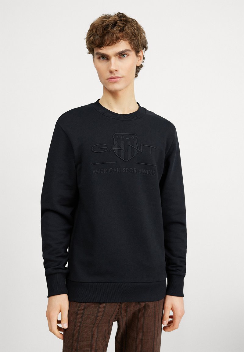 Fekete pulóver kerek nyakkal, kiemelkedő GANT logóval és "American Sportswear" felirattal. Simább, puha anyagból készült.