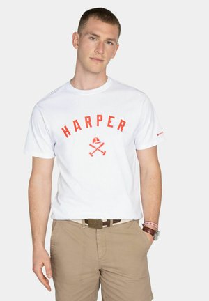 Joven con camiseta blanca "HARPER" con letras rojas y pantalones cortos beige, de pie con una mano en el bolsillo contra un fondo liso.