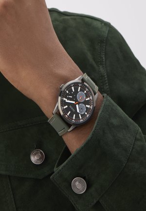 Analog-Uhr mit schwarzem Ziffernblatt, grünem Silikonarmband und silberfarbigem Gehäuse. Verfügt über auffällige Stundenmarkierungen und einen strukturierten Jackenkragen.