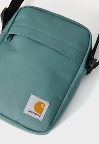 Light teal shoulder bag made of textured fabric, featuring a front pocket with a black zipper and a white label with an orange logo.