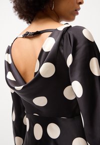 Rochie din satin negru cu buline mari crem, având un decolteu adânc în spate și închidere cu curea pe lateral, evidențiind o textură netedă și lucioasă.