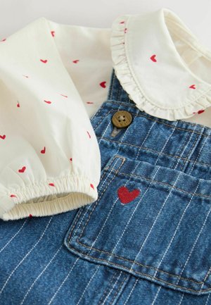 Jeanslatzhosen mit einem blau-weißen Streifenmuster, ausgestattet mit einer weißen Bluse mit roten Herzmustern und einem rüschigen Kragen. Rote Herzstickerei auf der Tasche.